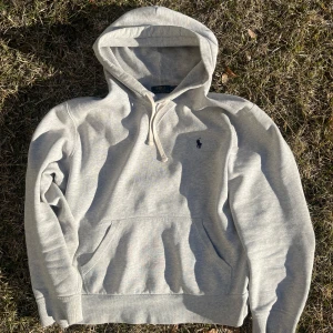 Ralph Lauren Hoodie - Säljer en klassisk grå hoodie från Ralph Lauren med justerbar dragsko i huvan och en broderad logotyp på bröstet. Perfekt för en avslappnad stil. Den har en stor ficka framtill och är gjord i mjukt material för extra komfort. Litet hål på baksidan, tror dock att det går att laga! Storlek M passar perfek på mig som är 180 och väger 80kg! Passar nog på dom flesta som bär M, hör av er vid frågor!