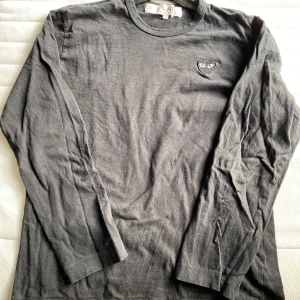 Svart långärmad tröja från Comme des Garçons - Säljer en svart långärmad tröja från Comme des Garçons med det ikoniska hjärta med ögon broderat på bröstet. Perfekt för en stilren och minimalistisk look. Tröjan är i storlek L och har en normal passform.