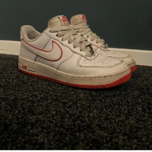 Nike Air Force med röda detaljer - Säljer ett par klassiska Nike Air Force med vit bas och röda detaljer. Skorna har en röd sula och röd Nike-logga på sidan. Perfekta för en sportig look. . 