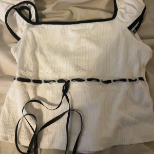 Vit ribbad topp med svarta detaljer - Säljer en söt vit ribbad topp med svarta kanter och knytband. Toppen har spetsdetaljer och är ärmlös med breda axelband. Perfekt för en somrig look!