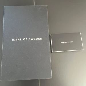 Svart mobilskal med kortfack från Ideal of Sweden - Snyggt svart mobilskal från Ideal of Sweden med praktiskt kortfack. Skalet har stilrena nitar och en metallplatta med logotyp. Perfekt för att hålla dina kort säkra och lättillgängliga.