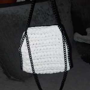 Vit virkad väska med svart kedja - Snygg vit virkas väska med svarta kedjehandtag. Perfekt för att ge en unik touch till din outfit. Väskan har en enkel och stilren design som passar till många tillfällen.