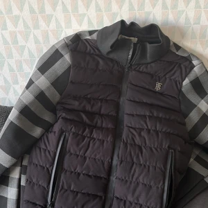 Svart Burrbyrr  jacka med rutiga ärmar - Snygg svart quiltad jacka med dragkedja och rutiga långärmade ärmar. Perfekt för en stilren look med en touch av mönster. Jackan har en liten broderad detalj på bröstet. Storlek S