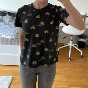 Svart t-shirt med regnbågsmönster från KappAhl - Snygg svart t-shirt från KappAhl med ett coolt regnbågsmönster i brunt och beige. Perfekt för en avslappnad stil. T-shirten har korta ärmar och en rund halsringning.
