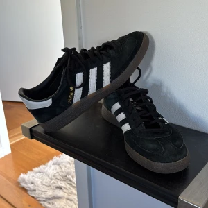 Svarta Adidas gazelle spezial  - Säljer ett par svarta Adidas sneakers med vita detaljer och klassiska tre ränder på sidan. Skorna har en snygg mockayta och snörning framtill. Perfekta för en stilren look.