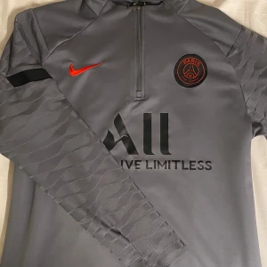 Psg hoodie - Snygg grå långärmad tröja från Nike med Paris Saint-Germain-logga och röd Nike-swoosh. Tröjan har en halv dragkedja och ett subtilt mönster på ärmarna. Perfekt för fotbollsfans!