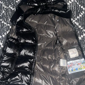 Svart dunjacka från Moncler - Snygg svart dunjacka från Moncler med glansig finish. Jackan har en dragkedja framtill och är quiltad för extra värme. Perfekt för kyligare dagar med stil.