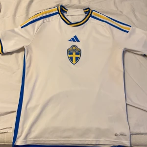 Vit fotbollströja från Adidas - Säljer en vit fotbollströja från Adidas med Sveriges landslagsemblem på bröstet. Tröjan har blå och gula detaljer samt korta ärmar. Namnet 'ISAK' och nummer 9 är tryckt på baksidan. Perfekt för fotbollsfans!