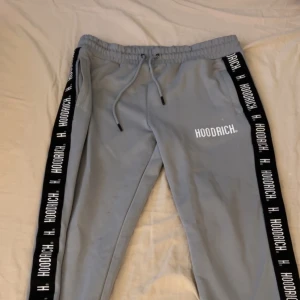 Grå mjukisbyxor från Hoodrich - Snygga grå mjukisbyxor från Hoodrich med svart och vit logotyp längs sidorna. Byxorna har en elastisk midja med snörning för justerbar passform. Perfekta för en avslappnad stil.