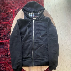 Svart Burberry Hoodie - Burberry Zip Up Hoodie, svart storlek M. Ett av snörena är trasiga men resten är i toppskick, som ny. Pris kan diskuteras lite.