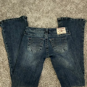 Blå jeans från True Religion - Snygga blå jeans från True Religion med klassisk femficksdesign och märkets ikoniska sömmar på bakfickorna. Jeansen har en bootcut-stil och låg midja, perfekt för en avslappnad look.