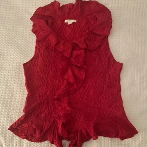 Röd ärmlös blus med volangdetaljer - Säljer en elegant röd blus med volangdetaljer och krage. Blusen är ärmlös och har en fin struktur i tyget som ger en unik look. Perfekt för att piffa upp garderoben! Jag säljer den för jag tar inte på mig, jag har haft den på en gång så den är ny skick