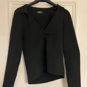 Svart långärmad topp från ONLY - Snygg svart topp från ONLY med lång ärm och v-ringad design. Toppen har en elegant knytning framtill och en klassisk krage. Perfekt för en stilren look.