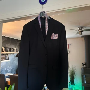 Mörkblå kostym från Selected Homme - Elegant mörkblå kostym från Selected Homme endast använd en gång och är perfekt för formella tillfällen. Kavajen har en klassisk design med två knappar och en bröstficka perfekt för en näsduk och är i storlek 54. Byxorna har en rak passform och är tillverkade i ett bekvämt material och är i storlek 52. Slipsen och näsduken ingår. Skriv för fler frågor, bilder och prisförslag.