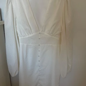 Vit klänning från NA-KD - Elegant vit klänning från NA-KD med v-ringning och långa ärmar. Klänningen har knappar framtill och ett diskret prickigt mönster som ger en stilren look. Endast använd en gång i ett fåtal timmar. Nypris 600kr, kan gå ned i pris vid snabb affär💗