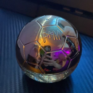 BParfym i fotbollsinspirerad flaska - BUnik parfymflaska formad som en fotboll med en glänsande, metallisk yta. Flaskan har en spegelblank finish som reflekterar ljus i olika färger. Perfekt för sportentusiaster som vill ha en doft med stil. Flaskan är både dekorativ och funktionell med en praktisk sprayfunktion.