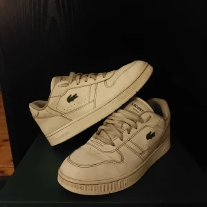 Vita sneakers från LacosteVita sneakers från Lacoste - Snygga vita sneakers från Lacoste med klassisk design och snörning. Skorna har en diskret krokodil-logga på sidan och är perfekta för en stilren look. De är tillverkade i syntetmaterial och har en bekväm passform.