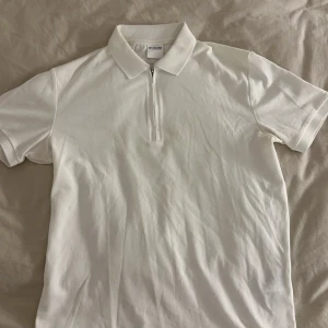 Vit Selected Homme t-shirt med zip - Vit t-shirt från Selected Homme med klassisk pikékrage och half zip framtill. T-shirten har korta ärmar och är gjord i mjuk bomull. Kragen har en snygg meshstruktur som ger extra stilpoäng. Perfekt för dig som gillar clean och fräsch look.