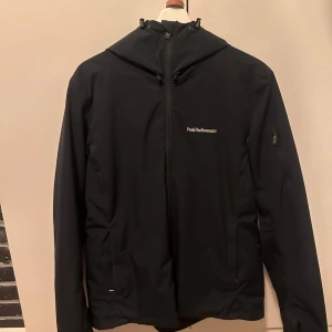 Svart vindjacka Peak Performance - Snygg svart vindjacka från Peak Performance med stilren design och diskret logga på bröstet. Jackan har huva med snörning, hel dragkedja framtill och praktiska fickor med dragkedja. Perfekt för dig som gillar sportig och clean look.
