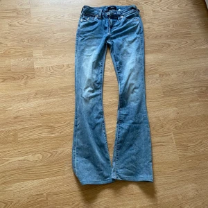 True Religion Becca Bootcut Jeans - Snygga blå jeans från True Religion, modell Becca Mid Rise Bootcut. Klassisk femficksdesign, coola detaljer på bakfickorna och silverfärgade knappar. Jeansen har en bootcut passform och är tillverkade i mjukt denimtyg. Perfekta för dig som gillar en avslappnad men trendig look. En liten fläck som knappt syns.