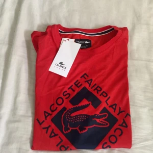 Röd Lacoste Sport t-shirt med tryck - Röd t-shirt från Lacoste Sport med stort mörkblått tryck framtill och klassisk rund hals. Snygg detalj med randig insida i halsen. Perfekt för dig som gillar sportig och stilren design.