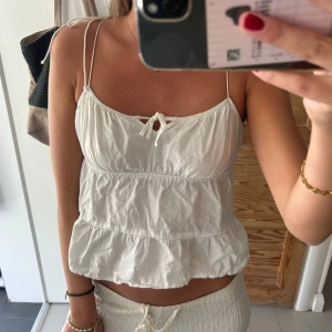 Vit volang linnetopp med smala band - Supersöt vit linnetopp med tunna axelband och volangkant. Toppen har en liten knytning framtill och är croppad med luftig passform. Perfekt för varma dagar och ger en chill vibe till din outfit.