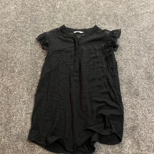 Svart blus med spets från H&M - Svart blus från H&M med snygga spetsdetaljer över axlar och rygg. Blusen har korta ärmar med volang och en liten knapp framtill vid halsen. Materialet känns lätt och luftigt, perfekt för dig som gillar en stilren look med lite extra edge. Fått sååå mycket komplimanger i denna😍