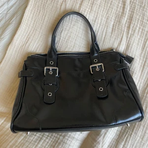 Svart handväska med silverdetaljer - Snygg svart handväska i skinn med två rejäla handtag och coola spänndetaljer i silver. Väskan har dragkedja upptill och metallfötter undertill som ger extra edge. Insidan är rymlig med fack för småsaker. Perfekt storlek för allt du behöver ha med dig.