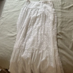 Vit långkjol med volanger - Säljer en vit långkjol med bred resår i midjan och fina volangdetaljer. Kjolen har ett subtilt mönster i tyget som ger en extra touch. Perfekt för dig som gillar en romantisk och luftig stil.