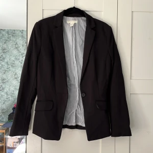 Kavaj H&M - Svart damkavaj med vit/grå-randigt innertyg. Två fickor på framsidan. En knapp på framsidan. Storlek: 36. Material utsida: 76% polyester, 19% viskos, 5% elastan. Material insida: 100% polyester. I använt skick, men ingen slitning. Lite missfärgad på märkeslappen i nacken. 
