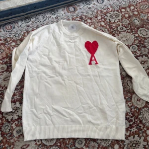 Ami Paris ny  - Säljer en vit stickad tröja från Ami Paris med ett stort rött hjärta och bokstaven A broderat på bröstet. Tröjan har rund hals, långa ärmar och ribbade muddar vid ärmslut och nederkant. Perfekt för dig som gillar stilrena detaljer och vill sticka ut.