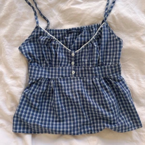 Rutig babydoll top från Brandy Melville - Supersöt blå och vit rutig linnetopp från Brandy Melville med smala axelband, v-ringning och knappar framtill. Toppen har en söt spetskant vid halsen och knytband i ryggen för extra passform. Perfekt för varma dagar och chill vibes.