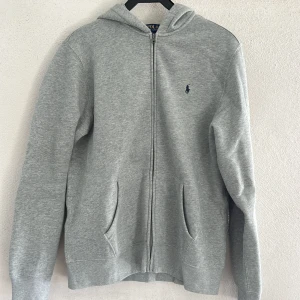 Grå hoodie från Polo Ralph Lauren XL som passar S/m - Snygg grå hoodie från Polo Ralph Lauren i storlek XL. Som passar S/M Tröjan har huva, dragkedja och två fickor framtill. Klassisk blå broderad logga på bröstet. Perfekt för chill dagar och enkel att matcha med jeans eller joggers. Aldrig använd!!😁