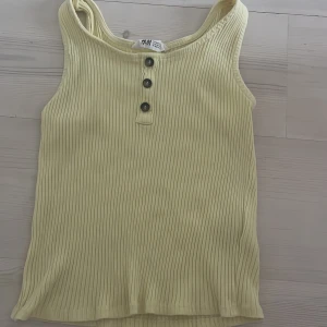 Ljusgul ribbad linnetopp från H&M - Snygg ljusgul linnetopp från H&M med ribbad struktur och tre svarta knappar framtill. Toppen är ärmlös och har en rundad hals. Perfekt för varma dagar och enkel att matcha med jeans eller shorts.