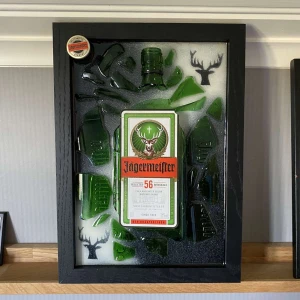 Jägermeister - Detta unika inramade konstverk handgjord av Epoxy Creations, visar en Jägermeister-flaska med detaljer och krossat glas, perfekt för dig som gillar kreativitet och Jäger. Passar som dekoration eller som present till någon som älskar Jäger! Gjort på återbruk✅