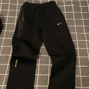Nike NOCTA svarta mjukisbyxor S - Svarta mjukisbyxor från Nike NOCTA med gul logga och detaljer. Byxorna har snörning i midjan, dragkedja på ena fickan och NOCTA-tryck på benet. Skön passform och stilren design, perfekt för streetwear och chill.