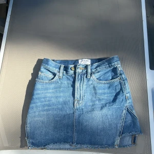 Blå jeanskjol från FRAME, strl 23 - Snygg kort jeanskjol från FRAME i klassisk blå denim. Kjolen har rå kant nertill, fyra fickor och bälteshällor. Perfekt för dig som gillar en chill och trendig look. Passar dig som vill ha en enkel men cool stil. ny pris är 3698kr