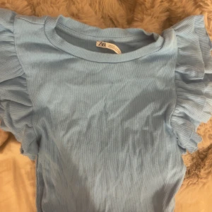 Ljusblå ribbad topp med volangärmar Zara - Supersöt ljusblå topp från Zara med ribbad struktur och volangdetaljer på ärmarna. Modellen är kortärmad och har en rund halsringning. Perfekt för dig som gillar en feminin och trendig stil.