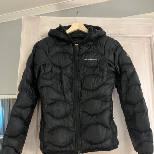 Svart dunjacka Peak Performance  - Svart Peak Performance Helium Jacka. Mycket bekväm jacka som inte passar mig längre. Tyvärr så har insidan på ena fickan gått sönder men går att sy igen den så funkar de, finns även några få små hål som är lappade på insidan i luvan, inget man tänker på. Utöver de är jackan i mycket gott skick! Storlek Xs, pris 600:-