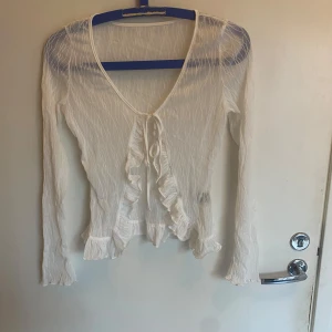 Vit omlottopp med volanger och knyt - Säljer en superfin vit omlottopp med lång ärm och v-ringning. Toppen har transparent, lätt ribbad struktur och snygga volangdetaljer längs kanten samt knyt framtill. Perfekt för dig som gillar romantisk och trendig stil.