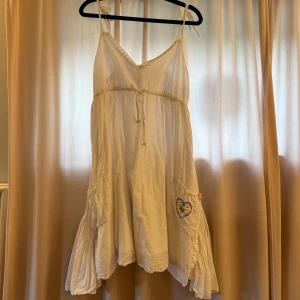 Vit klänning från Koola Anna - Supersöt vit klänning från Kooal Anna med tunna axelband, v-ringning och knyt i midjan i storlek M. Klänningen har volangdetaljer, fickor och en broderad hjärtformad patch på sidan. Tillverkad i linne material.✨✨