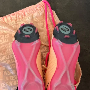 Nike Phantom rosa fotbollsskor - Nike Phantom fotbollsskor i ljusrosa med svarta detaljer och snörning. Skorna har en rund tå och platt sula med rosa och svarta dobbar för grymt grepp på planen. Ovandelen är i syntetmaterial med mönstrad struktur för bättre bollkontroll. Kommer med en matchande rosa Nike-påse. Skorna är använda 1 gång (se första bilden) 