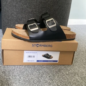 Svarta sandaler från Stormberg strl 40 - Stormberg Sandö Cork Sandal W i svart med två breda remmar och stora silvriga spännen. Snygg korksula och mjuk innersula för extra komfort. Perfekt för chill dagar och enkel att matcha med jeans eller shorts.