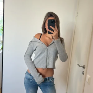Grå croppad hoodie med dragkedja - Säljer en grå croppad hoodie med dragkedja framtill och ribbad struktur. Tröjan har lång ärm, huva och offshoulder-design som ger en chill vibe. Perfekt att matcha med jeans eller shorts för en avslappnad look.