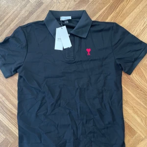 Svart piké från Ami med rött hjärta - Svart pikétröja från Ami Alexandre Mattiussi med klassisk krage och korta ärmar. På bröstet finns ett broderat rött hjärta med bokstaven A. Materialet är mjuk bomull och tröjan har diskret Ami-logga broderad vid nederkanten. Perfekt för en clean och stilren look.