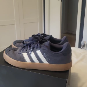 Adidas sneakers i mörkblå mocka - Snygga Adidas sneakers i mörkblå mocka med klassiska vita ränder på sidan och gummisula i beige. Skorna har rund tå, snörning och låg profil. Perfekt för dig som gillar stilrena och sportiga vibes. Ikonisk Adidas-logga på sidan och sula. Skorna är i storlek 40 och är mycket bra skick, priset kan sänkas lite vid snabbt köp