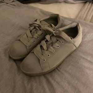 Gråa sneakers från din sko - Snygga gråa sneakers med breda matchande snören och platt sula. Skorna har en stilren design med silverdetalj på hälen och är tillverkade i ett mjukt syntetmaterial. Perfekta för dig som gillar minimalistisk stil och vill ha ett par neutrala sneakers till din garderob. Köpta på Din Sko, men märket är attitude.