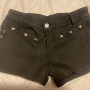 Svarta shorts med dödskalle-detaljer - Coola svarta shorts med snörning på sidorna och små silvriga dödskalle-detaljer vid fickorna. Modellen är tight och lowaist, med klassiska bakfickor och bälteshällor. Materialet känns som bomull med lite stretch för extra komfort.