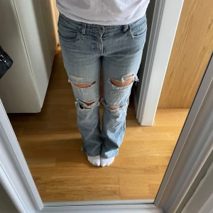 Ljusblå ripped bootcut jeans - Säljer ett par ljusblå jeans med vida ben och stora slitningar över knäna. Jeansen har klassisk femficksdesign, normal midja och är tillverkade i bomull. Perfekta för dig som gillar en avslappnad och trendig look. Har lagat dem en gång tid grenen och finns även ett hål på baksidan, men som inte stör vid användning