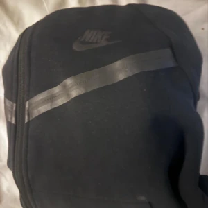 Svart Nike kofta med reflexdetaljer - Svart kofta från Nike med diskret logga på bröstet och snygga reflexdetaljer över bröstet. Koftan har dragkedja framtill och är perfekt för dig som vill ha en sportig och clean look. Passar till både chill och aktiva dagar.